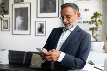Positive handsome caucasian senior businessman in suit and glasses typing on smartphone