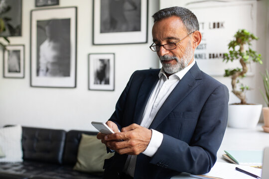 Positive Handsome Caucasian Senior Businessman In Suit And Glasses Typing On Smartphone