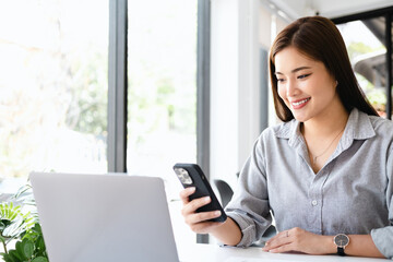 Cheerful woman entrepreneur sitting beside a window and smiling. Businesswoman working in office with a mobile phone in hand.
