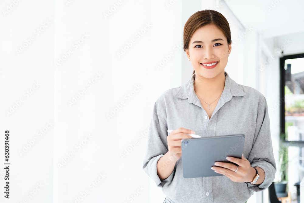 Wall mural young asian business woman wearing shirt using tablet standing in modern workspace. smiling confiden