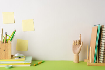 Creative inspiration scene. Side view photo stationery-filled desk with pencils, pens, books holder, notepads, wooden mannequin hand, stapler, sticky notes on beige wall backdrop. Ad space available - obrazy, fototapety, plakaty