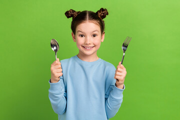 Photo of funny satisfied small schoolgirl dressed blue sweatshirt hold spoon fork ready for dinner...