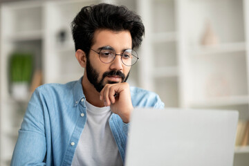 Thoughtful Young Indian Man In Glasses Sitting At Workplace And Looking At Laptop Screen