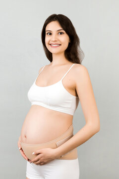 Portrait of pregnant woman in underwear wearing maternity belt to reduce pain in the back at gray background with copy space. Orthopedic abdominal support belt concept