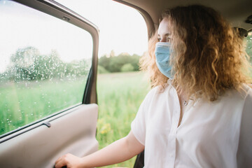 Stylish young carly woman in medical mask looking out window while sitting on back seat of car on blurred background with sunset