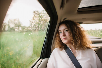 Cheerful positive curly young woman in casual wear sitting in automobile backseat with fastened seatbelt and looking away.