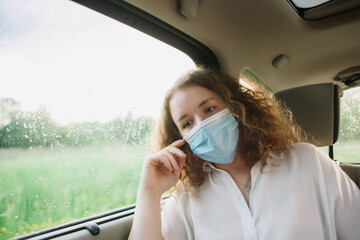 Stylish young carly woman in medical mask sitting on back seat of car on blurred background with sunset.