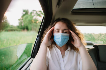 Stylish young carly woman in medical mask have a headache while sitting on back seat of car on natural background with sunset