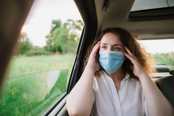 Stylish young carly woman in medical mask have a headache while sitting on back seat of car on natural background with sunset