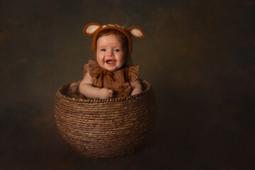 portrait of smiling happy beautiful baby girl in felted deer bonnet hat sitting in basket wearing brown dress with bow four month milestone