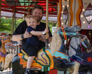 happy little toddler boy riding colorful carousel horse at carnival festival fair together with his smiling dad family fun holiday weekend