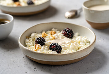 Cottage cheese in ceramic plates with blackberries, cream or yogurt, nuts and cup of coffee aside over grey concrete background