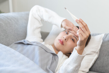 Sick, influenza asian young woman, girl have a fever, hand holding thermometer, touching forehead for check measure body temperature, illness while lying rest on sofa at home. Health care concept.