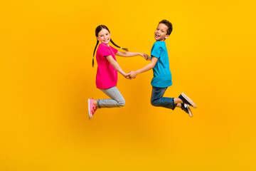 Fototapeta na wymiar Full size profile portrait of two carefree cute kids jumping hold arms rejoice isolated on yellow color background