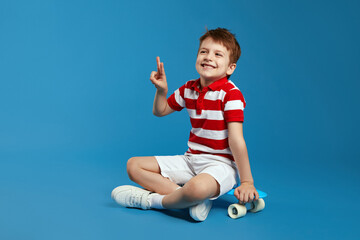 Excited little boy in trendy red striped polo shirt smiling and showing peace gesture while sitting on modern skateboard against blue background