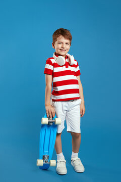 Vertical photo of full body stylish kid boy wearing red striped polo shirt and headphones on neck, leaning on skateboard and smiling for camera against blue background