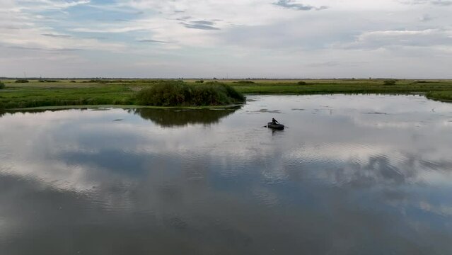 A man with fishing poles rides a boat through the steppe river where the water mirrors the sky.