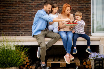 Family with a mother, father, son and daughter sitting outside on steps of a front porch of a brick house and eating strawberries
