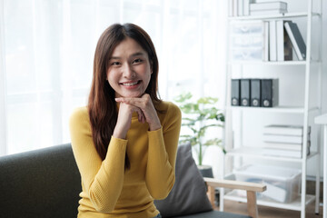 Young beautiful Asian woman in yellow sweater sitting on the sofa, relaxing in her living room at home.