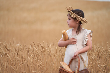 A cute little girl in a light linen dress with a wreath of ears on her head holds a bread basket in one hand and a long bread in the other hand against a golden background of a wheat field.