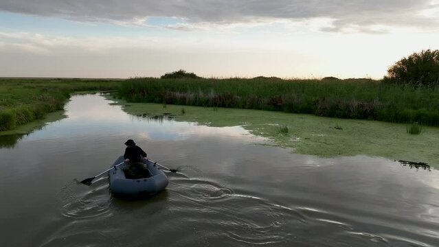 A man with fishing poles rides a boat through the narrow river in the calm, cloudy evening.