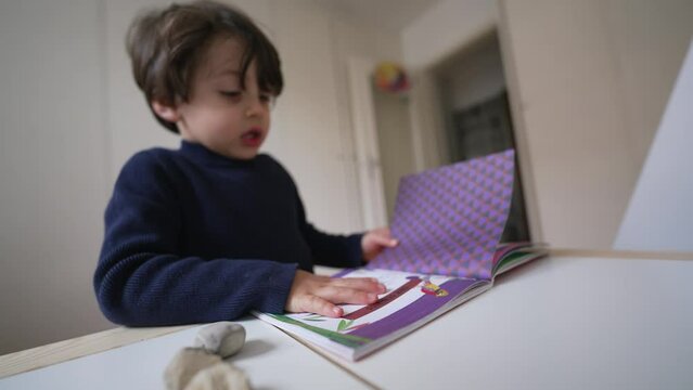 Child turning book pages looking at images and text. Little boy reading kindergarten school material