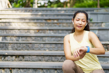 Happy cheerful Asian female runner sitting on the stair at the park after have a small running, woman using fitness tracker to track her exercise or workout progress.