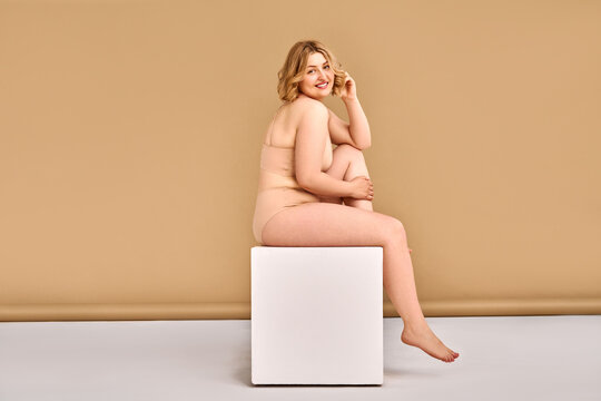Body positivity, confidence and love for yourself and your body. Attractive adult blonde plus size woman in beige lingerie sits on a cube in a photo studio on a beige background.