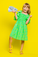 Rich pleased young cute school girl waving money dollar cash banknotes bills like a fan, success, lottery winner, big income, wealth. Teen blonde female child kid on studio yellow background. Vertical