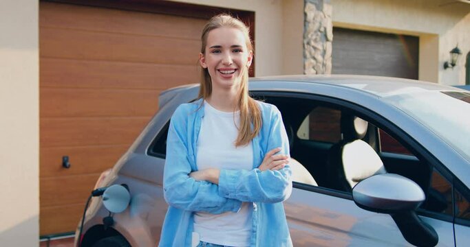 Portrait of young woman standing near parked car in the parking looking at camera smiling. Girl charging electric car at charging station near home.