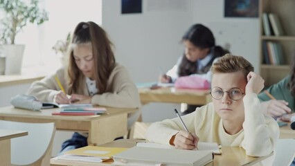 Selective focus of tired nerd boy sitting at desk in classroom making notes then looking at camera during lesson