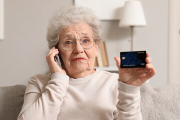 Shocked senior woman with credit card talking by mobile phone on sofa at home, closeup