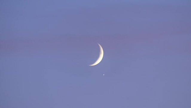 Blue sky and slowly moving pink clouds, crescent Moon and the light of the planet Jupiter. The conjunction of Jupiter with the Moon observed in October 2019.