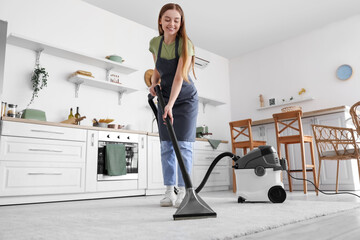 Young woman vacuuming carpet in kitchen - obrazy, fototapety, plakaty