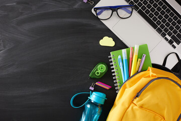Building the base for successful learning. Top view shot of laptop, goods for the school, schoolbag, eyeglasses, water bottle on blackboard background with empty space for promo or text