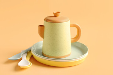 Teapot with plates and spoons for baby on pale orange background