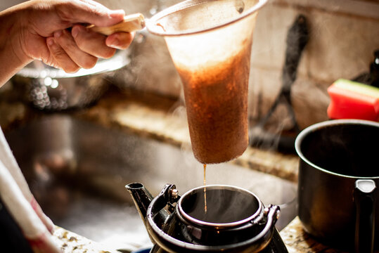 Mão coando café em um coador de pano em uma cozinha rústica, transmitindo a simplicidade e o sabor autêntico do café feito na hora.