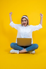 Woman doing winner gesture, full size body view woman sit floor ground legs crossed holding notebook on her lap clenched fists up air. Wearing casual white shirt and jeans. Studio yellow background.