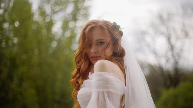 Portrait of a beautiful luxury fashion bride with red hair flowing in the wind. Attractive woman in a wedding dress with a veil gazes looking enchantingly into the camera.