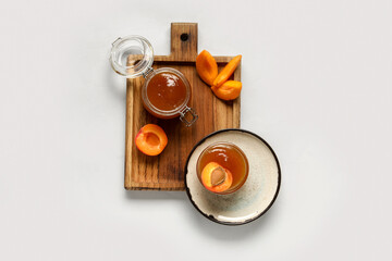 Wooden board with glass bowl and jar of sweet apricot jam on white background