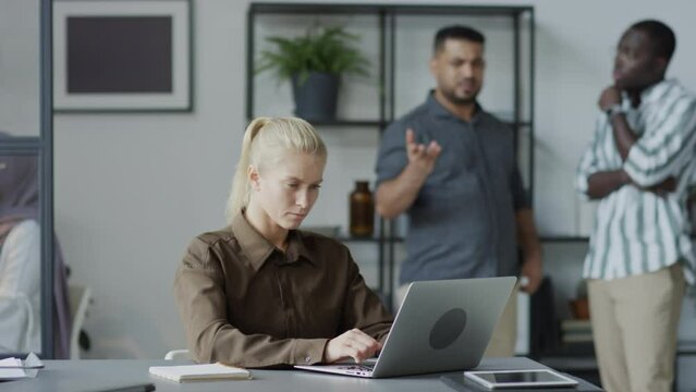 Selective focus shot of Caucasian female manager working on laptop trying to ignore annoying male co-workers slandering her