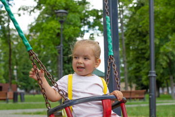 Fototapeta na wymiar A cute little girl swings on a swing in the park in summer and laughs. The child has fun in the summer in the park swinging on a swing. Happy child concept.
