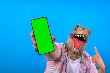 man with blurry dinosaur mask showing cell phone with green screen on isolated blue background