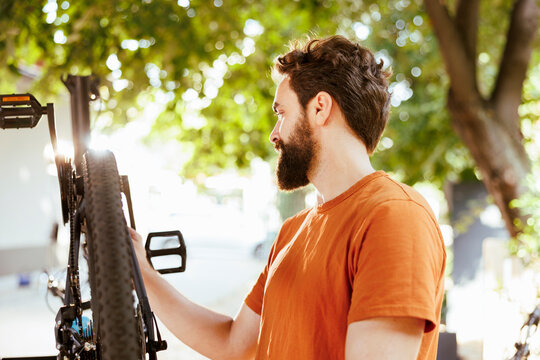 Detailed image showcasing young active caucasian man meticulously examining modern bicycle in home yard. Sporty male cyclist seen upkeeping and maintaining bike components outside.