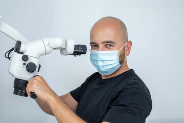 Male dentist in medical mask standing sideways near white dental microscope and holding hands on...