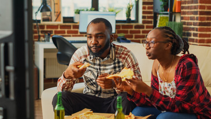 Happy life partners eating slices of pizza on couch, having fun together watching favorite movie on television. Young man and woman in relationship enjoying delivery food and alcohol.