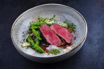 Gourmet barbecue dry aged angus beef steak with green asparagus and parmesan cheese in truffle mousse parfait and wild cherry relish cream served as close-up in a Nordic design bowl
