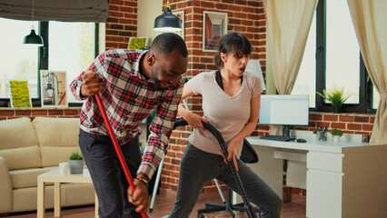 Diverse couple showing dance moves and having fun cleaning apartment, using mop to sweep dust and vacuum at home. Life partners dancing and singing, using washing solution, spring cleaning.