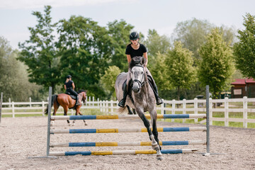 Girl rider on dapple gray horse jumping over triple bars in outdoor arena on a sunny summer day....