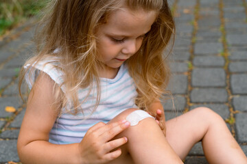 A little girl puts a band-aid on a wound on her knee after an accident. Selective focus.